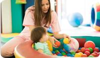 A child is in a play pen, while their occupational therapist models how to grab and hold a ball. One of the pediatric occupational therapy goals and objectives for this child has to do with strengthening their grasp on items, specifically being able to hold onto and eventually throw a ball.