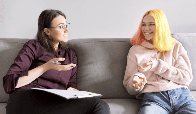 Two people sitting next to each other on a couch, speaking about ways a therapist can support their clients' coming out.