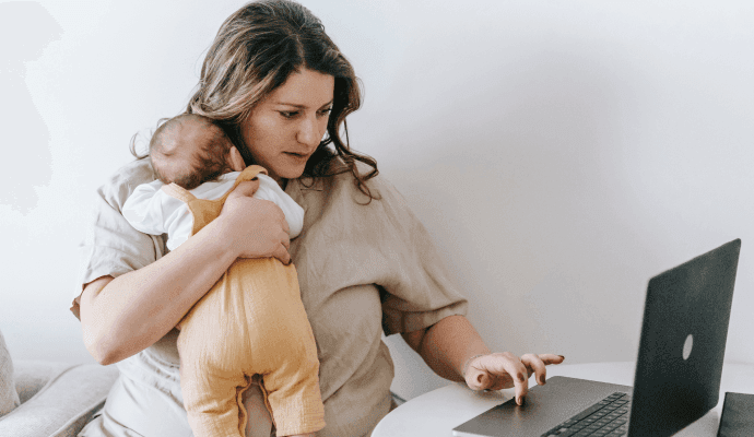 A woman sits holding a baby to her chest and looking at a laptop.