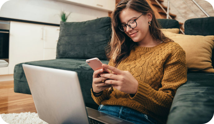 Woman smiling while looking at her phone with a laptop on her lap.